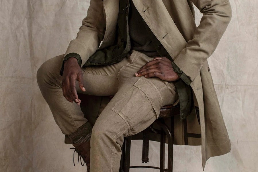 Beige trench coat with khaki pants and dark shirt seated on a wooden stool.