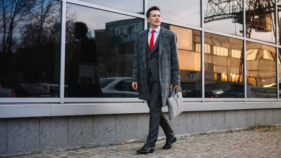 Businessman in grey overcoat and red tie walking on sidewalk for trench coat style tips.