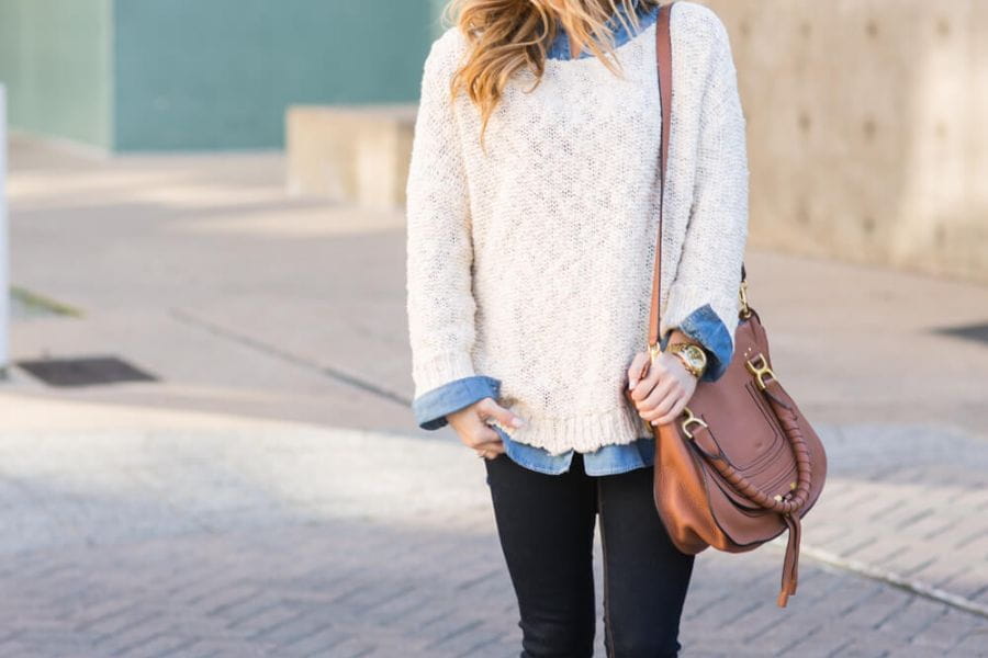 Cream-colored knit sweater layered over a blue shirt, paired with dark jeans and a brown bag.