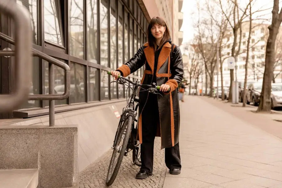 Cyclist in black and orange trench coat poses with bicycle on a stylish sidewalk.