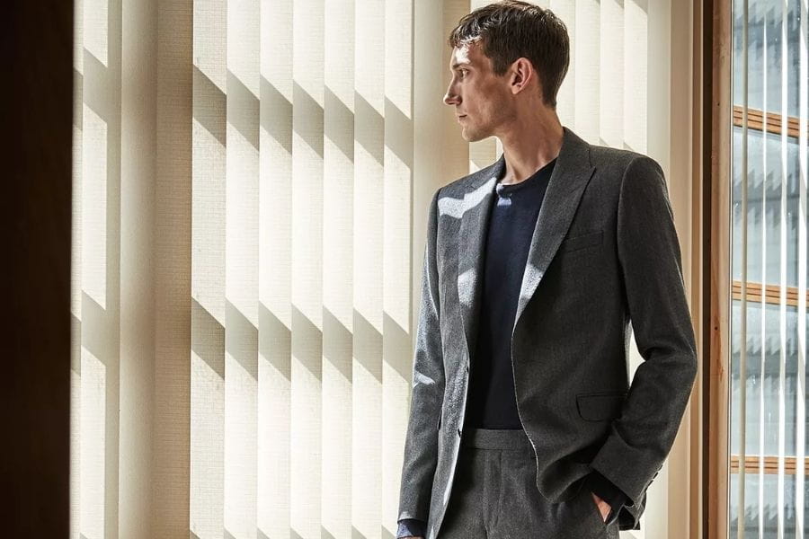 Man in gray suit jacket by window with Venetian blinds in men’s fashion guide.