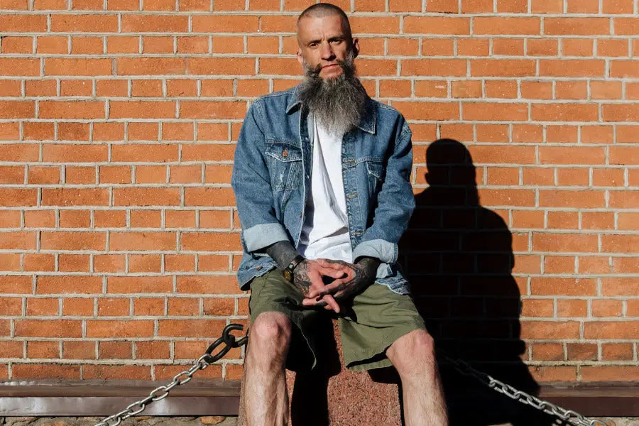 Person in a denim jacket and cargo shorts seated on concrete block by a brick wall.