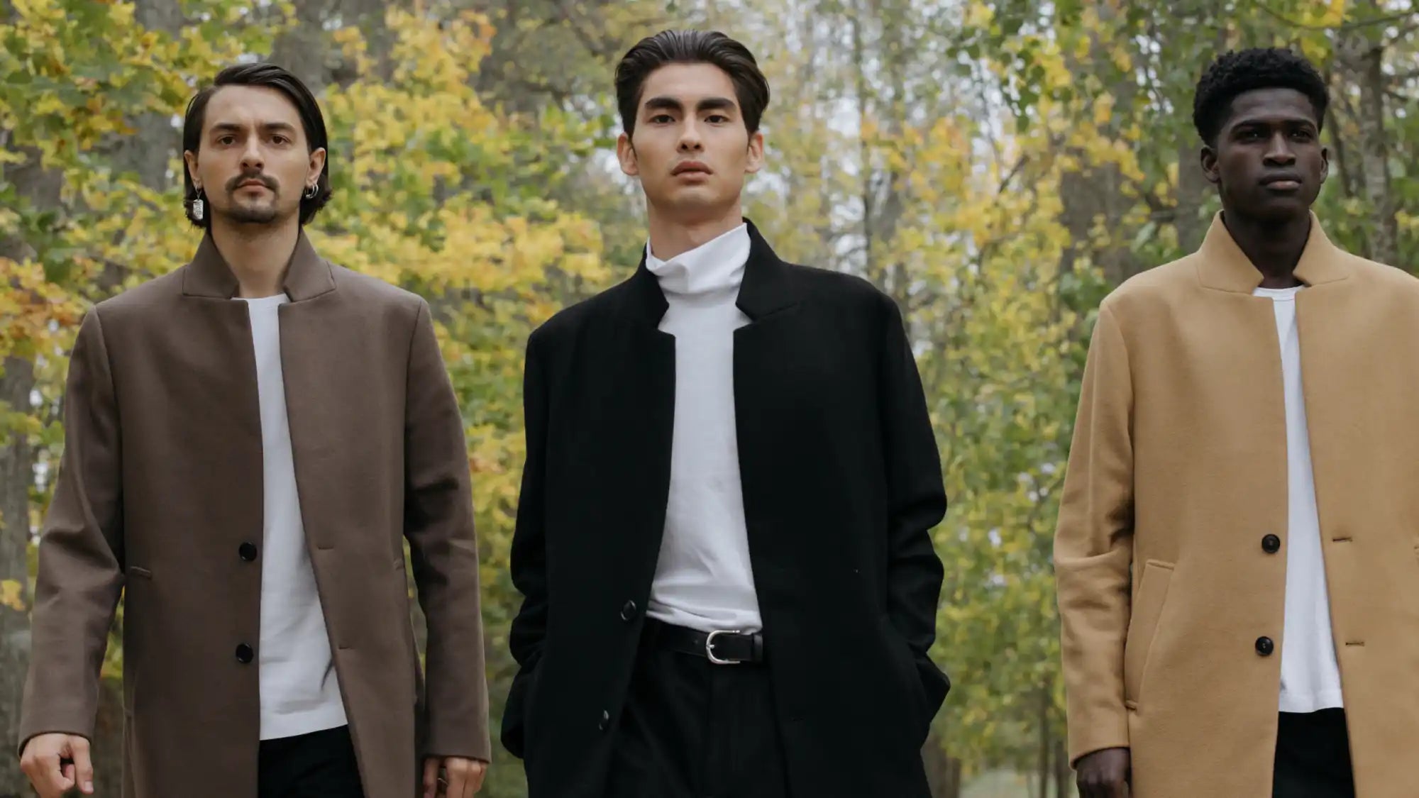 Three men standing side by side wearing stylish coats and turtleneck sweaters in an autumn setting.