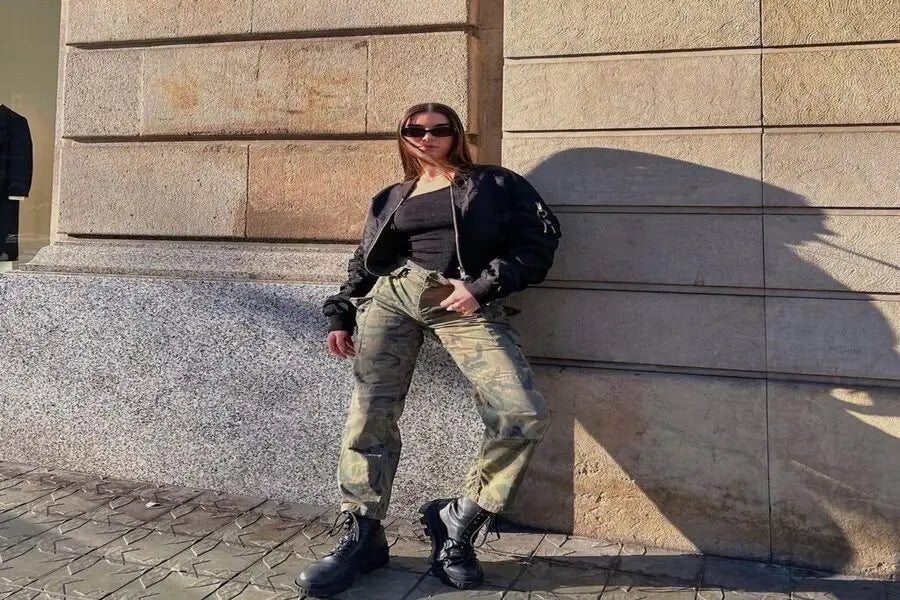 Person in cargo pants, black jacket, sunglasses, and chunky boots leaning against a wall.