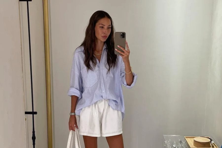 Woman in a light blue oversized shirt and white shorts taking a mirror selfie.