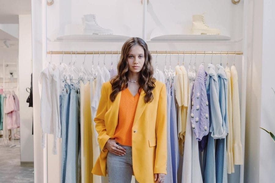 Woman in a yellow coat over an orange top and jeans at a clothing store.