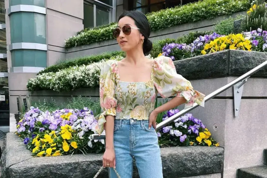 Woman in floral top and sunglasses poses by colorful flowers for summer outfit ideas.