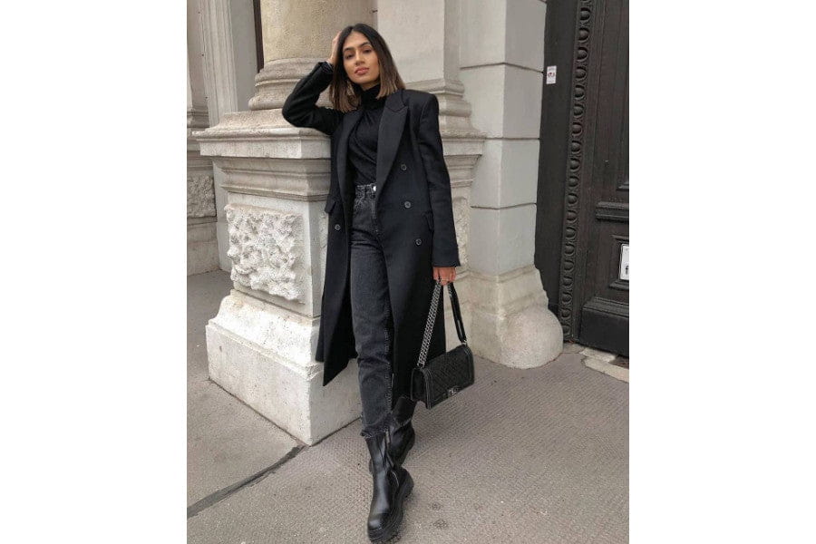 Woman in a long dark coat and boots showcasing minimalist apparel against a building.