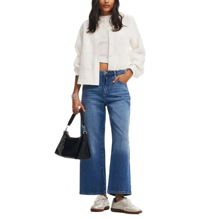 Stylish woman in Desigual jeans, white jacket, crop top, sneakers, and black handbag