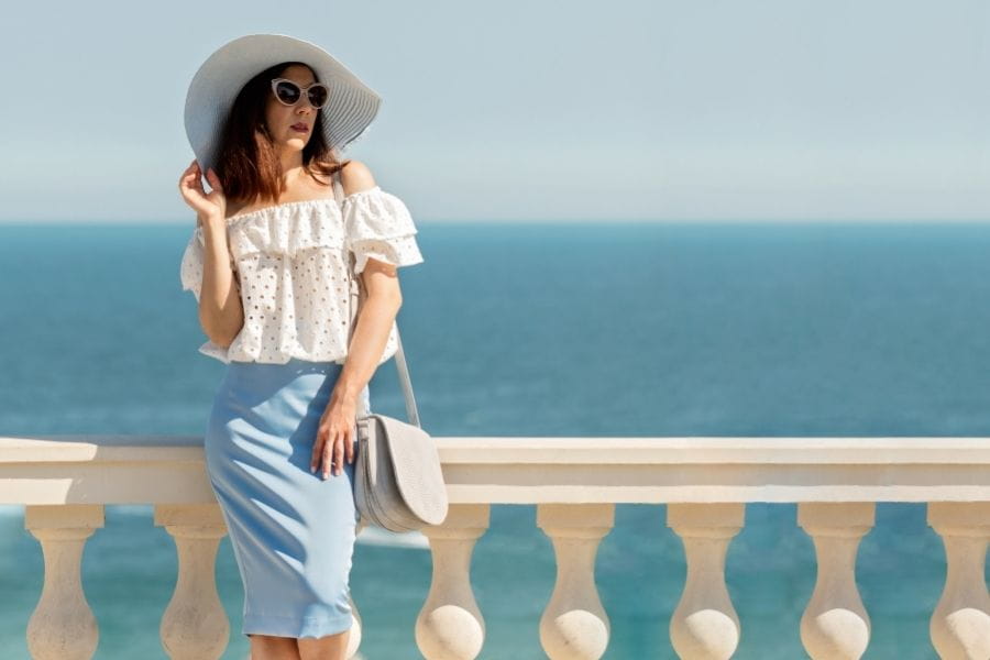 Woman in a white sun hat and off-shoulder top poses elegantly by a balustrade.